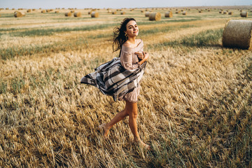 Naklejka na ściany i meble Full length portrait of a beautiful brunette in a dress and with a warm plaid. Woman enjoying a walk in a wheat field with hay bales