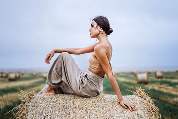 Barefoot brunette in linen pants and bare shoulders sitting on a hay bales in warm autumn day. Behind her is a wheat field © bedya