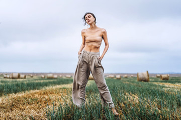 Brunette in fashionable clothes with bare shoulders standing in a wheat field on green grass. Woman holds her hands in pants pockets and looking forward
