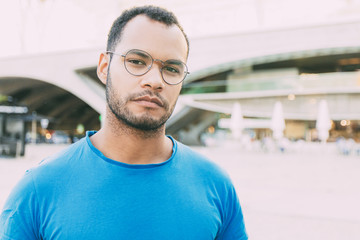 Fototapeta premium Serious confident man posing in city square. Front portrait on young Latin man wearing blue casual shirt and eyeglasses and looking at camera. Male portrait concept