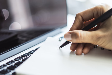 Woman writes with a pen in notebook on laptop keyboard in a sunny office, business and education...