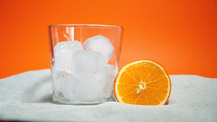 glass with ice and soda water and orange on sand on an orange background