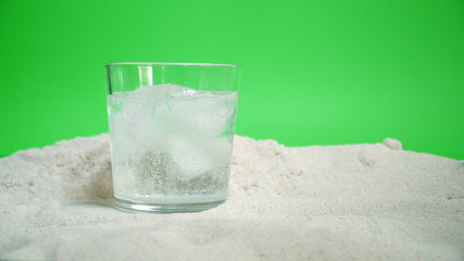 glass with ice and soda water on the sand on a green background