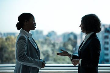Female business colleagues chatting in office corridor. Business women standing near office window...