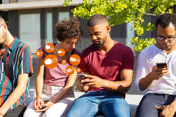 Cheerful handsome man showing phone screen to disgusted woman. People with smartphones and social media notifications icons sitting on parapet outdoors. Wireless connection concept