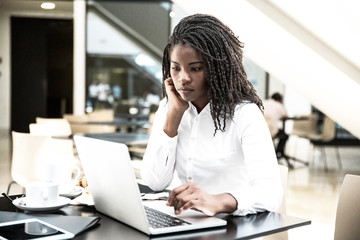 Fototapeta premium Focused young female office worker working in coffee shop. African American business woman using laptop in cafe. Wireless connection concept