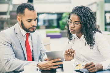 Serious business colleagues watching content on tablet together. Diverse business man and woman sitting in cafe, holding tablet and looking at screen. Media content concept