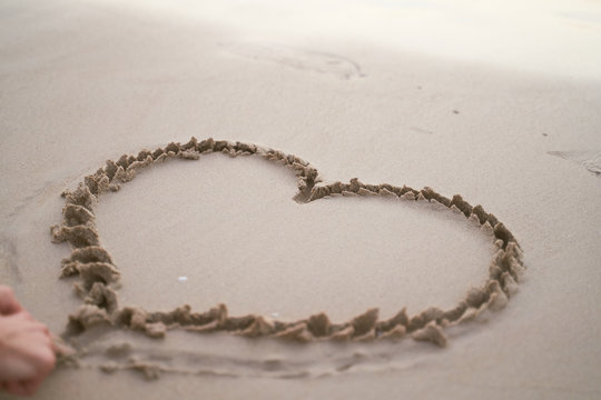 Drawing of heart on sand, waves of Andaman sea. Evening photo on Phuket Patong beach in Thailand. Love to travel. The girl draws or draws with fingers in sand.