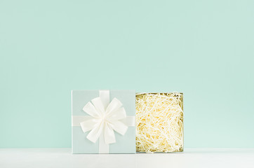 Festive background with opened standing empty square gift box with paper filler, ribbon, knot on soft light green mint menthe wall and white wood table. - Powered by Adobe