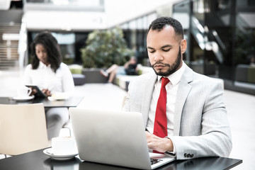 Serious focused male manager working on report and using laptop in coffee shop. Young black woman using tablet in background. Wireless technology concept