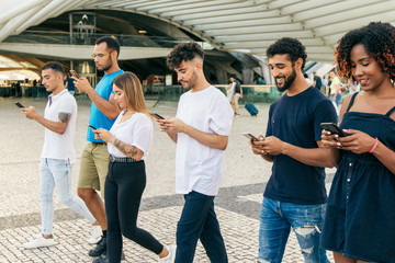 Multiethnic group of friends walking together and chatting online on smartphones. Line of men and women going outside and using mobile phones. Digital technology concept