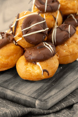 Profiteroles, coated in chocolate, stacked on a wooden chopping board with a blue tea towel.  On a wood background