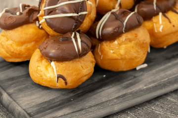 Profiteroles, coated in chocolate, stacked on a wooden chopping board with a blue tea towel.  On a wood background