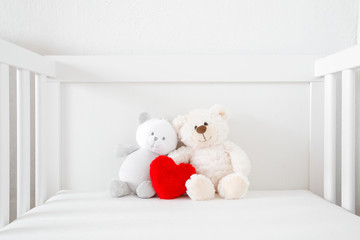Two smiling white teddy bears sitting in baby bed. Red fluffy heart in middle. Front view. Closeup.
