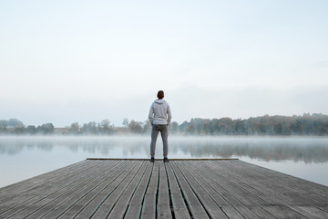 Young man standing alone on wooden footbridge and staring at lake. Thinking about life. Mist over water. Foggy air. Early chilly morning. Peaceful atmosphere in nature. Enjoying fresh air. Back view. - Powered by Adobe
