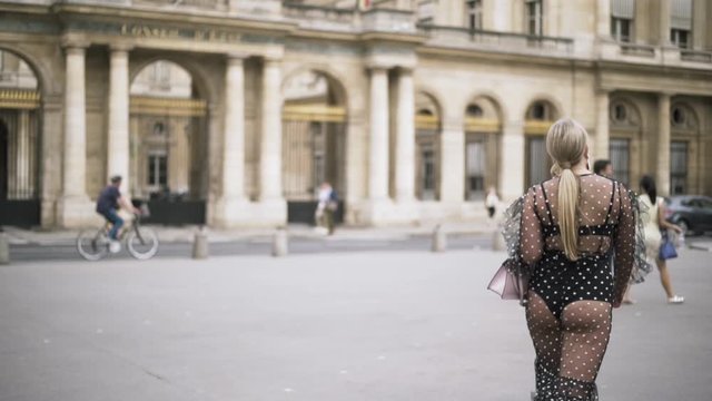 Young pretty woman with blond hair in front of a big historic building, France, travelling in Europe. Action. Sexy lady wearing chiffon dotted dress walking in the street.