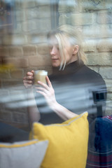 Attractive young blonde girl siting near window and drinking coffee or tea  in the cafe. Time to relax.