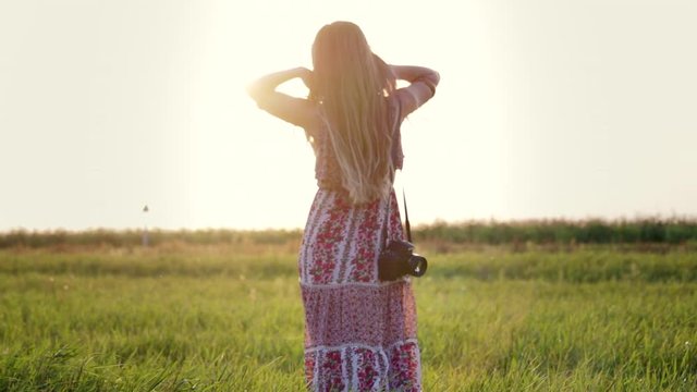 Rear view of a pretty young hippie woman with a camera on her shoulder walking through an agricultural field at sunset and touching her long hair. Girl in a sundress in nature, slow motion
