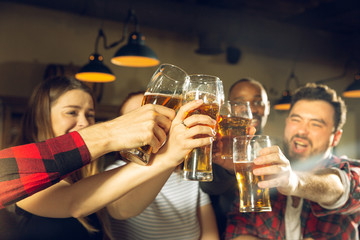 Sport fans cheering at bar, pub. Clinking beer glasses while watching championship, competition. Multiethnic group of friends excited in translation. Human emotions, expression, supporting concept.