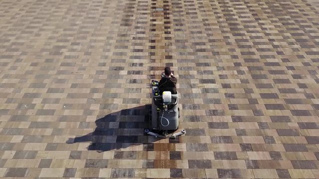 Aerial view of compact street sweeper car cleaning city sidewalk paving slabs with high pressure washers and rotating brooms outdoors. Road sweeping machine janitor leaves wet footprint on square.