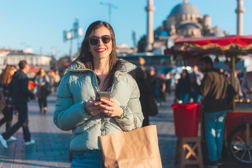 Obraz premium Cheerful brunette woman texting a message enjoying and laughing also keeps a shopping bag in a town center a near mosque
