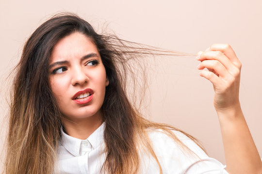 hair problems. young woman in white shirt checking her britle, damaged, and split hairs against pink background