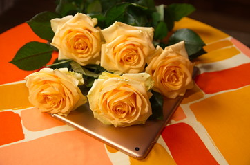  On a yellow-orange silk tablecloth lies a tablet and a bouquet of fresh beautiful fragrant roses.