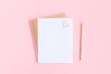 Blank white sheet on spiral golden notepad with paper clip heart and pencil on pink pastel...