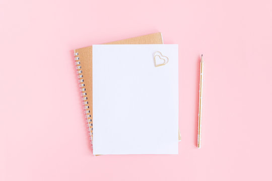 Blank white sheet on spiral golden notepad with paper clip heart and pencil on pink pastel background. Mock up for your text and design. Top view Flat lay Minimal style