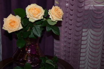 Against the background of purple textile, on a round glass table in a beautiful vase there is a bouquet of beautiful fresh fragrant orange roses with green leaves.