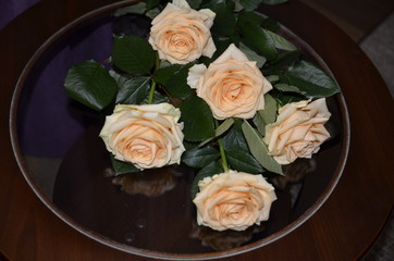  Against the background of purple textile, on a round glass table is a bouquet of beautiful fresh fragrant orange roses with green leaves.
