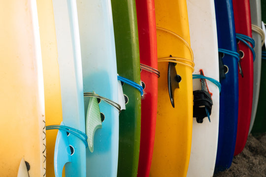 Close up Set of different color surf boards with fins in a stack on sandy beach for rent. Multicolored surfboards as rainbow.
