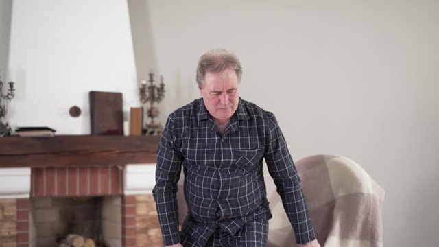 Positive mature Caucasian man standing up from chair and sitting down because of sudden sharp pain in back. Health problems of elderly retirees, aging concept, medical troubles.