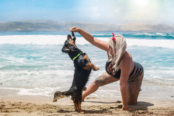 Young blonde girl in a black bikini, playing with her dog on the beach that is jumping to catch the stone that her owner is holding.