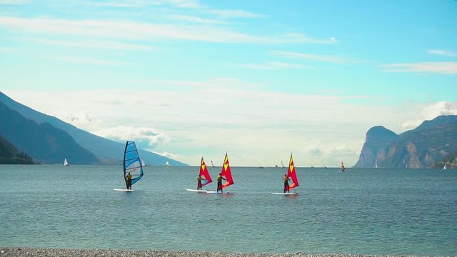 Riva Del Garda lake of Torbole Nago resort tourists engaged in water sports windsurf, rowin wave. Enjoy clean air wind spee extreme. Backdrop Italian alpine mountains sky blue. Lago di Garda Benaco