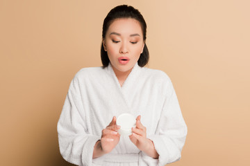 shocked beautiful asian woman in bathrobe holding cosmetic cream on beige background