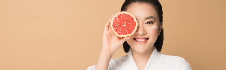 smiling beautiful asian woman in bathrobe with face cream on cheek and grapefruit half on beige...