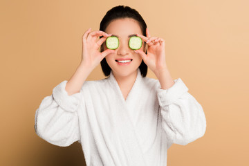 smiling beautiful asian woman in bathrobe with cucumber slices on eyes on beige background
