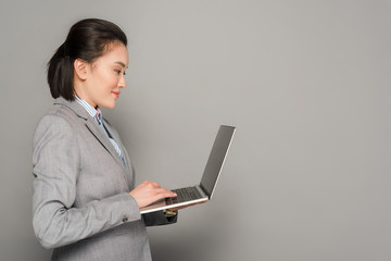 side view of smiling young businesswoman in suit using laptop on grey background