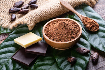 Cocoa powder in wooden bowl, bars of dark chocolate and cocoa butter with whole cacao beans  and fresh leaves on old rustic table.