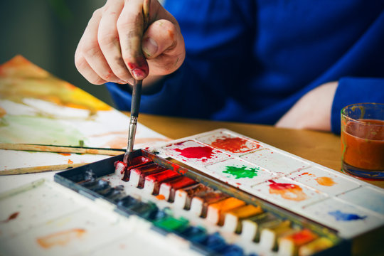 The artist holds a paintbrush with a dirty hand and scoops up a watercolor red paint from the palette, and next to it is a sheet with a sketch and a glass of dirty water for washing the brushes.