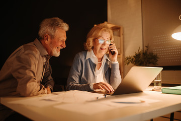 Portrait of blonde business woman working with laptop while having phone call late at night. Her partner is sitting next to her.