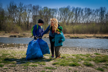 young blond hair woman with her sons in blue gloves cleaning the river bank from plastic and garbage in big blue trash bags on a sunny day in early spring