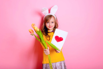 Happy Easter. Girl kid blonde in rabbit ears holding a card red heart, yellow tulips. Happy Mother's Day! International Women Day. Daughter's day.