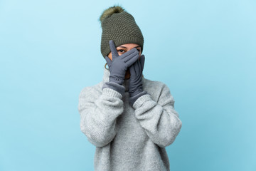 Young Uruguayan girl with winter hat isolated on blue background covering eyes and looking through...