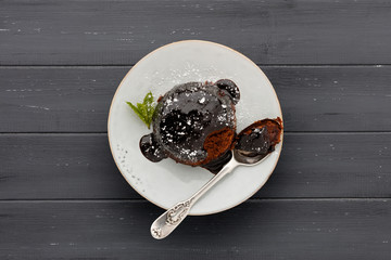 An individual chocolate sponge cake, with a sticky chocolate sauce, on a white plate with a spoon, shot on a dark grey wooden surface with copy space