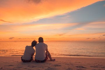 Couple in love watching sunset together on beach travel summer holidays. People silhouette from behind sitting enjoying view sunset sea on tropical destination vacation. Romantic couple on the beach - Powered by Adobe