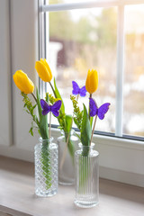 Composition with yellow vivid fresh tulips and violet butterfly shaped candies in glass vases, selective focus