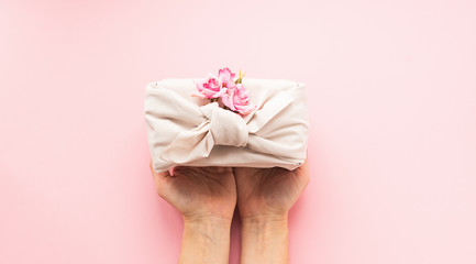 Female hands holding a holiday gift packed fabric in the manner of Furoshiki on a fashionable pink background.
