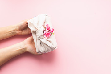 Female hands holding a holiday gift packed fabric in the manner of Furoshiki on a fashionable pink background.